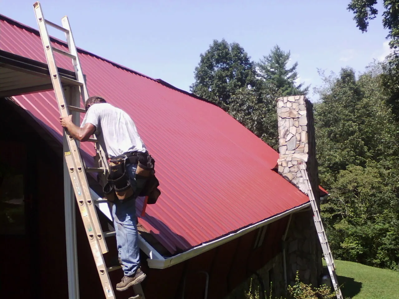 Roofer on a ladder beside a red metal roof and stone chimney — Soffit & Fascia Repair by Roofing Brothers in Millsboro